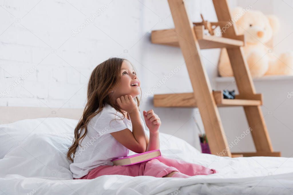 dreamy girl in pajamas looking up while sitting on bed with book