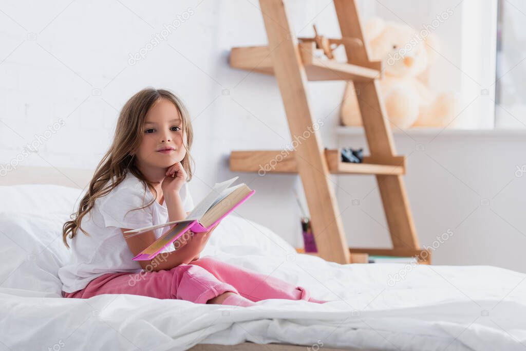 child in pajamas looking at camera while sitting on bed and reading book