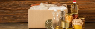 panoramic shot of cardboard box with groats near water, oil, canned food and honey on wooden background, charity concept clipart