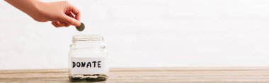 cropped view of woman putting coin in penny jar with donate lettering on wooden surface on white background, panoramic shot clipart