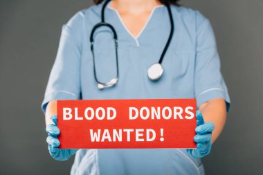 cropped view of doctor with stereoscope holding red card with blood donors wanted lettering isolated on grey clipart