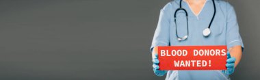 cropped view of doctor with stereoscope holding red card with blood donors wanted lettering isolated on grey, panoramic shot clipart