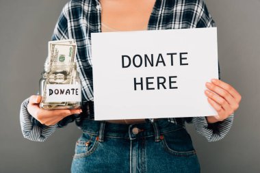 cropped view of woman holding card with donate here lettering and jar with money on grey background clipart