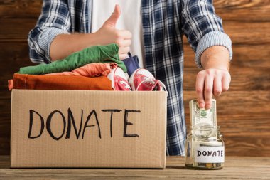 cropped view of man showing thumb up near cardboard box with donated clothes and money on wooden background, charity concept clipart
