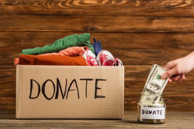 cropped view of man putting money in jar near cardboard box with donate lettering and clothes on wooden background, charity concept clipart