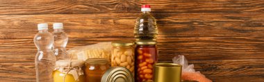 panoramic shot of groats near water, oil, canned food and honey on wooden background, charity concept clipart
