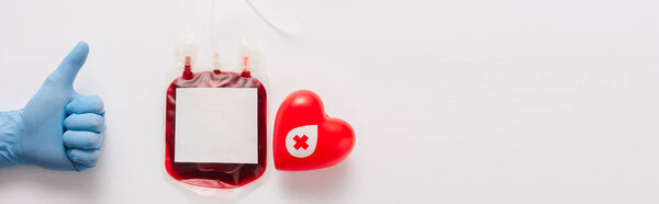 обрезанный вид врача, показывающего большой палец вверх рядом с пакетом крови и красным сердцем на белом фоне, панорамный снимок