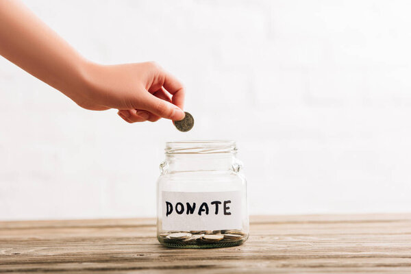 cropped view of woman putting coin in penny jar with donate lettering on wooden surface on white background