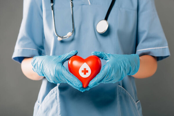 cropped view of doctor with stereoscope holding red heart isolated on grey, blood donation concept