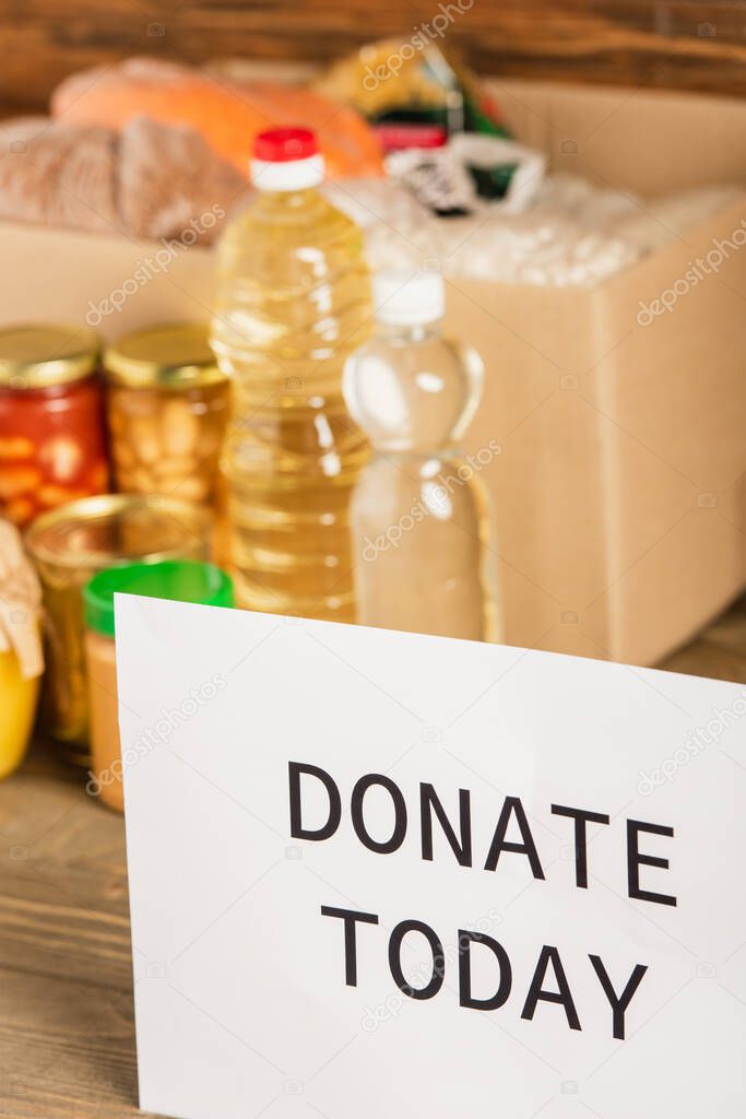 selective focus of cardboard box with food and donate today card on wooden background, charity concept