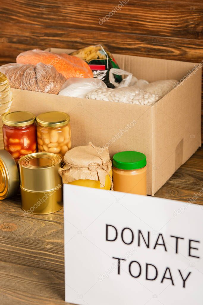 selective focus of cardboard box with food and donate today card on wooden background, charity concept