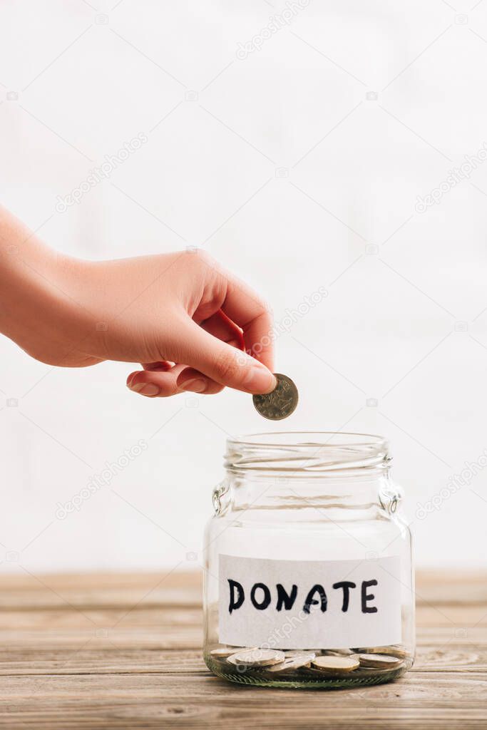cropped view of woman putting coin in penny jar with donate lettering on wooden surface on white background