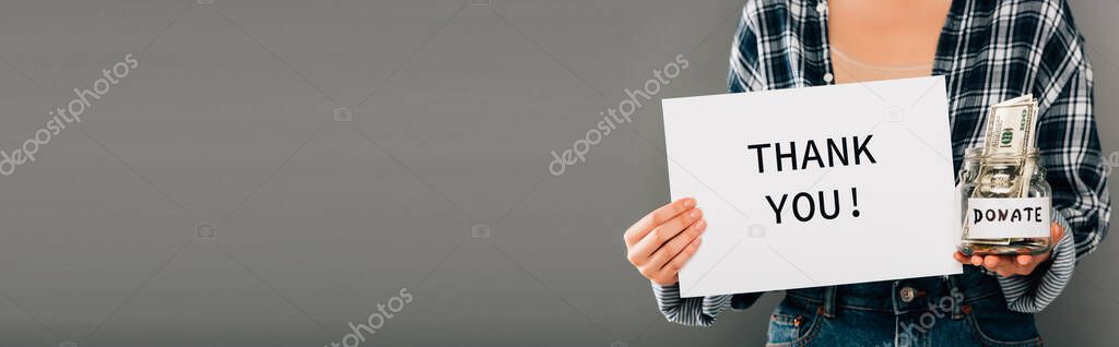 cropped view of woman holding card with thank you lettering and jar with donated money on grey background, panoramic shot