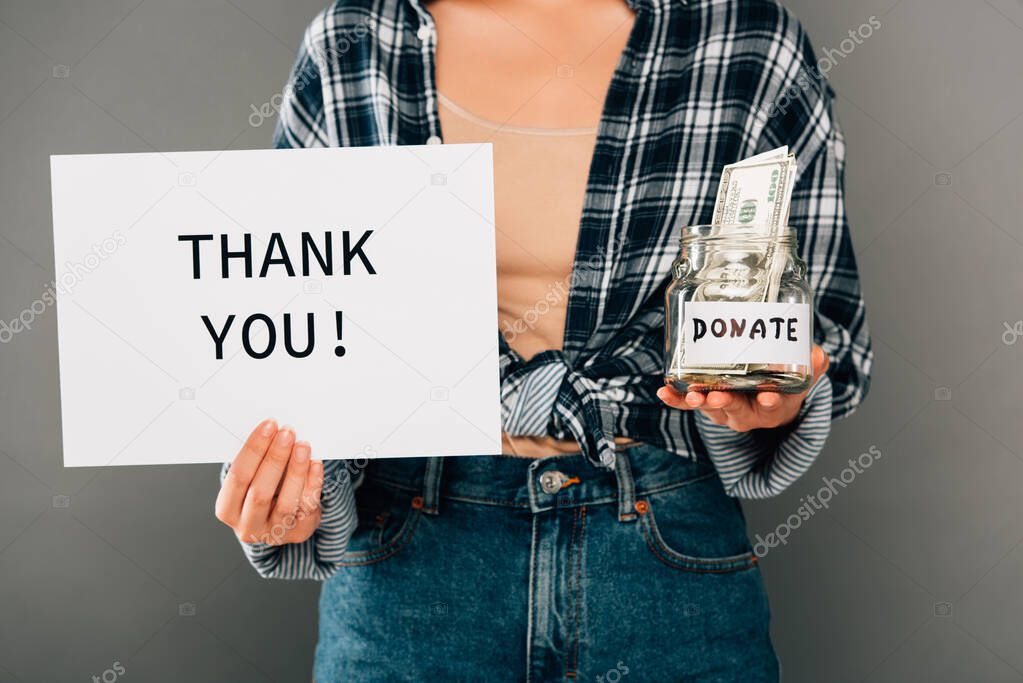 cropped view of woman holding card with thank you lettering and jar with donated money on grey background