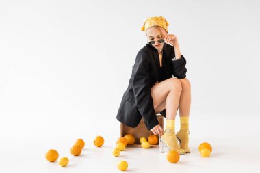 Beyaz arka planda dağınık narenciye meyvelerinin yanında ahşap kutuda poz veren şık bir kadın.