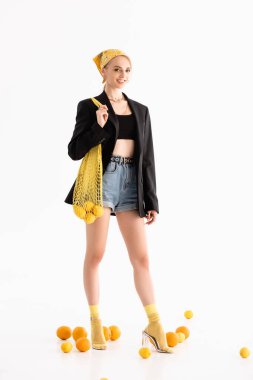 Beyaz arka planda dağınık narenciye meyvelerinin yanında sicim torbasıyla poz veren şık bir kadın.