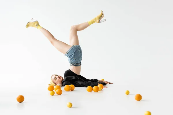 白い背景に柑橘系の果実が散在して足を上げて床にポーズをとるファッショナブルな女性 — ストック写真