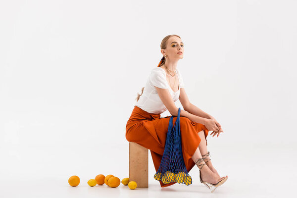 elegant blonde woman posing with string bag near scattered citrus fruits on white background