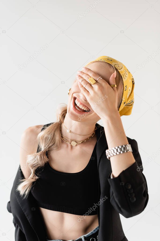 fashionable woman in yellow headscarf and golden accessories covering face with hand and laughing isolated on white
