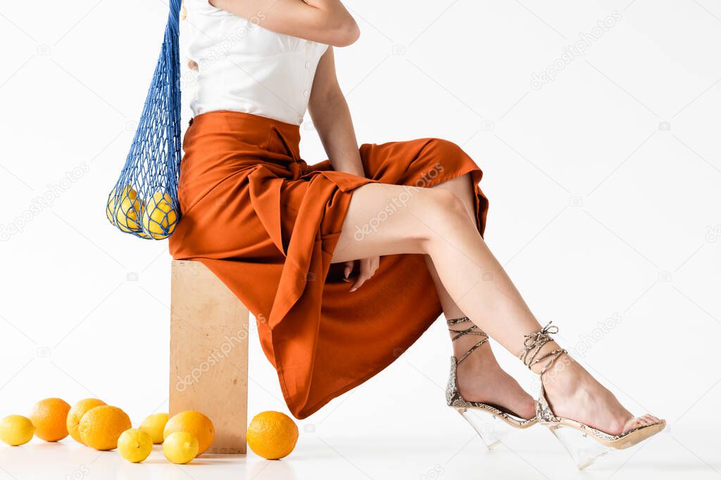 cropped view of elegant woman posing with string bag near scattered citrus fruits on white background