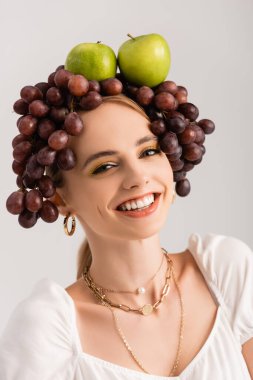 Kafasında elma ve üzümlerle poz veren kırsal sarışın kadın portresi beyaz üzerinde izole edilmiş.