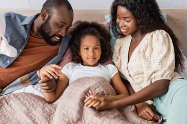 high angle view of african american girl lying in bed and looking at camera while parents sitting near and touching her hands clipart