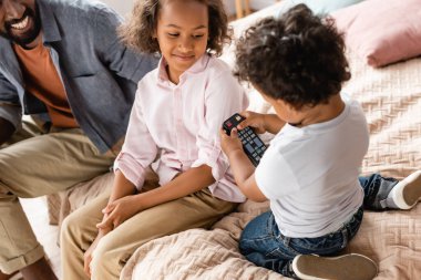 cropped view of african american man and girl sitting on bed near boy holding tv remote controller clipart