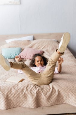 excited african american girl lying on bed with raised legs while having fun in bedroom clipart