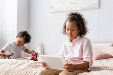 Erkek kardeşinin yanında dijital tablet kullanan konsantre Afro-Amerikan kız, yatak odasında oyuncaklarla oynuyor.