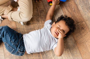 top view of sleepy african american boy touching eyes while lying on floor at home clipart