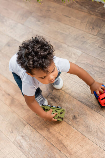 overhead view of curly african american child playing with toy vehicles on wooden floor