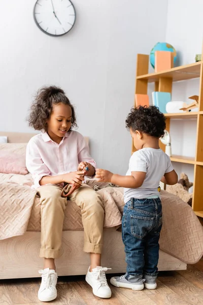 身穿休闲装的非洲裔美国女孩坐在床上 把玩具抱在小弟弟身边 — 图库照片