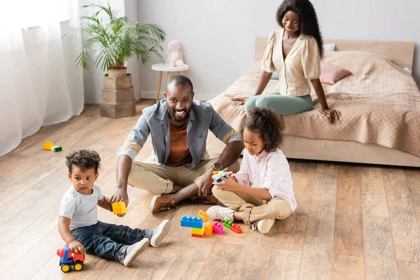 african american children and dad playing on floor with building blocks near mom sitting on bed