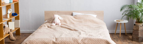 horizontal image of bedroom, bed with beige blanket, rack with books and potted plant 