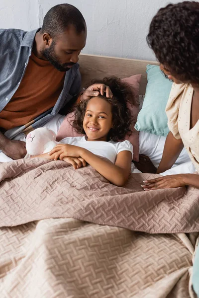 african american parents sitting near daughter lying in bed under blanket