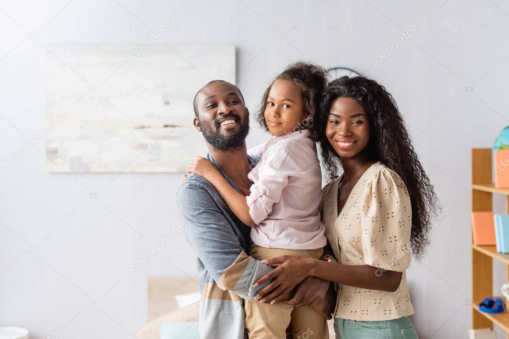 Bearded african american man holding daughter near mother hugging them and looking at camera