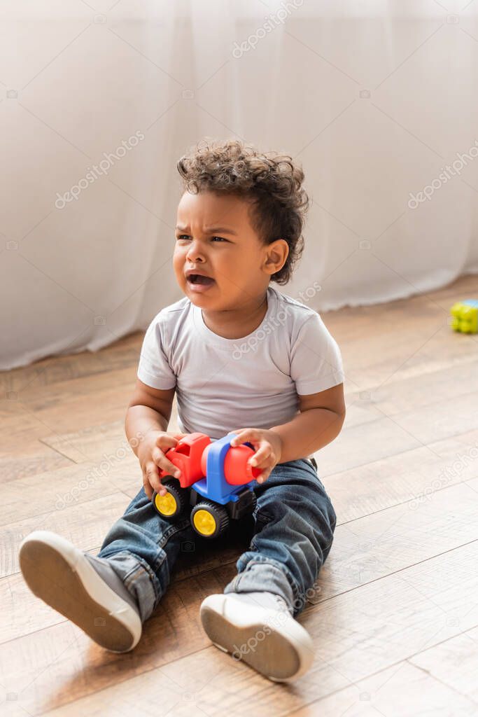 crying african american boy in white t-shirt and jeans sitting on wooden floor and holding toy truck