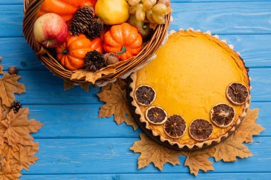 top view of autumnal harvest in wicker basket near pumpkin pie on blue wooden background clipart