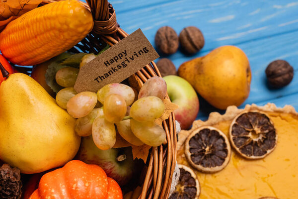 close up view of autumn harvest in wicker basket with happy thanksgiving card near pumpkin pie on blue wooden background