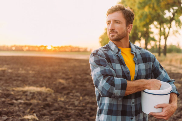 rancher in plaid shirt holding bucket while standing on plowed field in sunshine