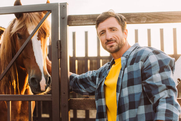 rancher in checkered shirt touching horse in corral and looking at camera