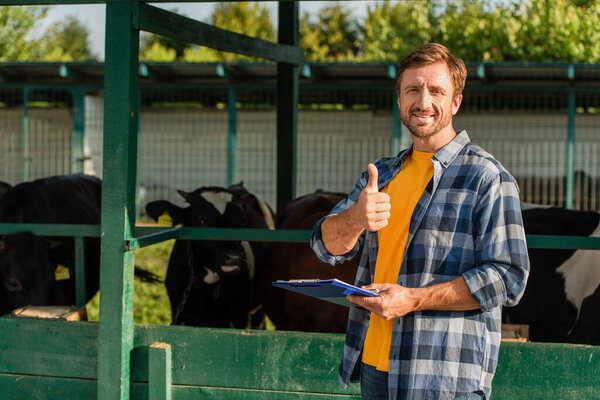 фермер в клетчатой рубашке показывает большой палец вверх, стоя рядом с коровником с планшетом