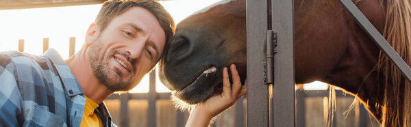 horizontal image of farmer looking at camera while touching horse, panoramic shot