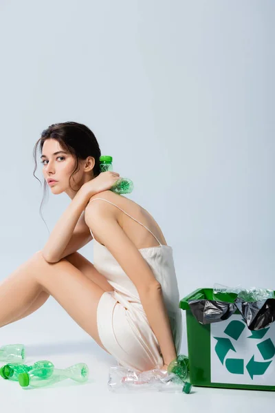プラスチックボトルとゴミ箱の近くに座ってシルクのドレスでブルネットの女性白 生態学の概念上のリサイクルサインと — ストック写真