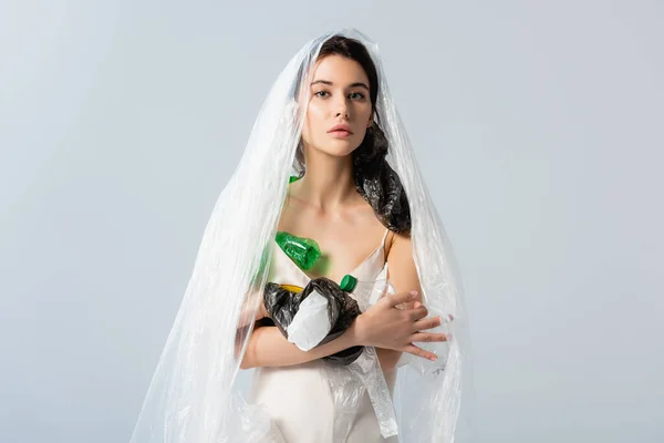 young woman with plastic bag on head holding empty bottles and looking at camera isolated on grey, ecology concept