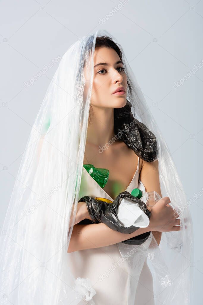 brunette woman with plastic bag on head standing in silk dress with empty bottles isolated on white, ecology concept  