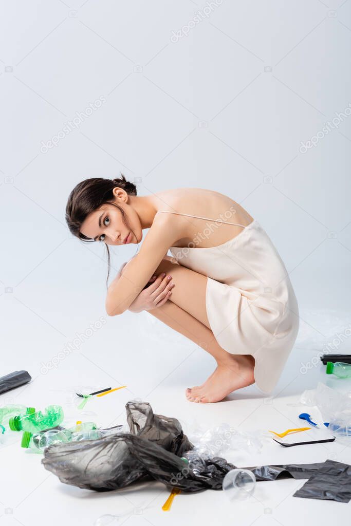 barefoot woman in silk dress looking at camera and sitting near plastic bags and bottles on white, ecology concept  