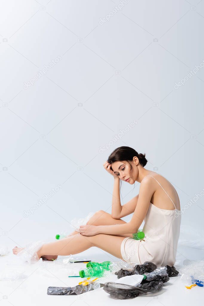 barefoot model in silk dress looking at camera and sitting near plastic bags and bottles on white, ecology concept  