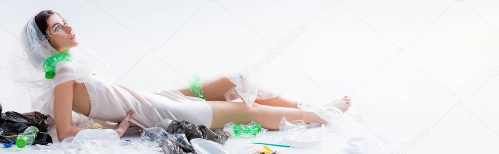 horizontal image of barefoot woman in silk dress with plastic bag on head sitting near empty bottles on white, ecology concept  