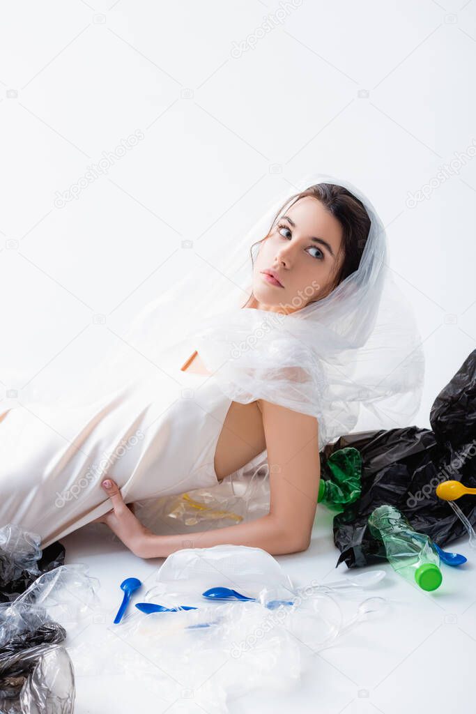 woman in silk dress with plastic bag on head sitting near empty bottles and looking away on white, ecology concept  